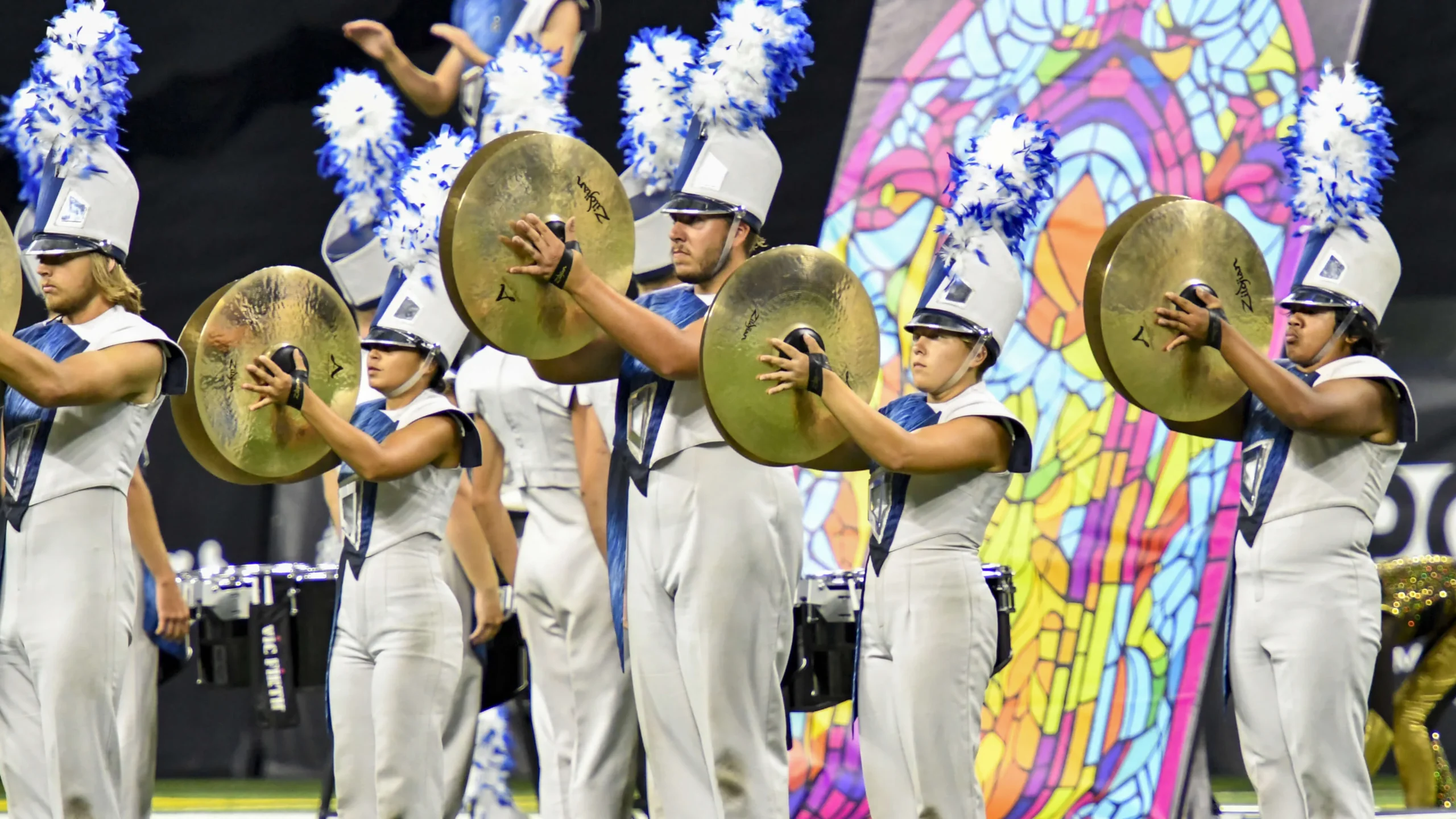 A diverse group in white and blue uniforms energetically playing drums in a vibrant setting